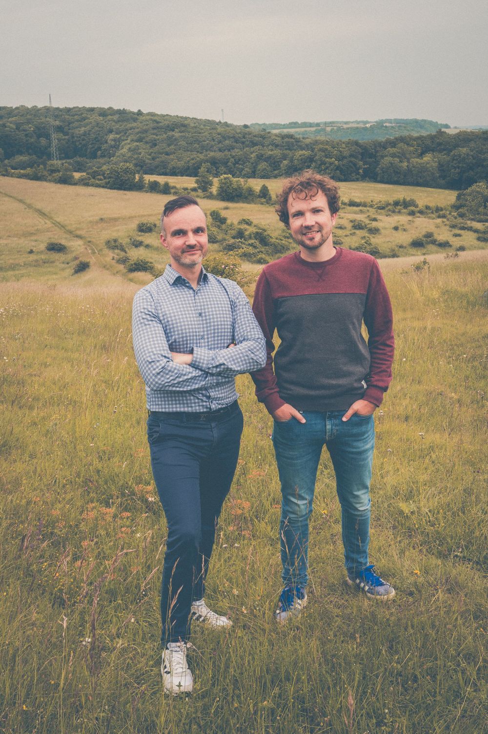 Two people stand together in an autumn field with rolling hills and trees in the background.