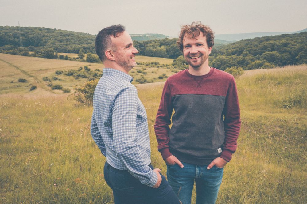 Two people standing and smiling in a sunny field with rolling green hills in the background.