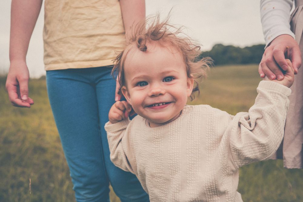 A toddler wearing a beige sweater smiles joyfully while walking outdoors with adults on a sunny day.