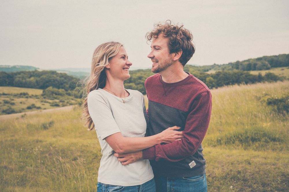 A couple embraces while standing together in a scenic green meadow on a sunny afternoon.