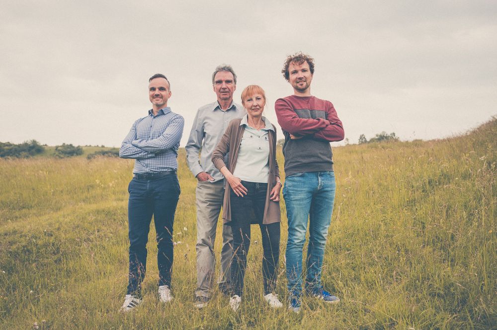 Four people stand together in casual attire on a grassy hillside on an overcast day.
