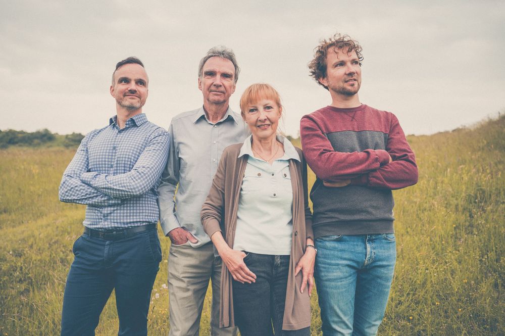 Family group standing together outdoors in a grassy field with mountains in background, wearing casual clothing.