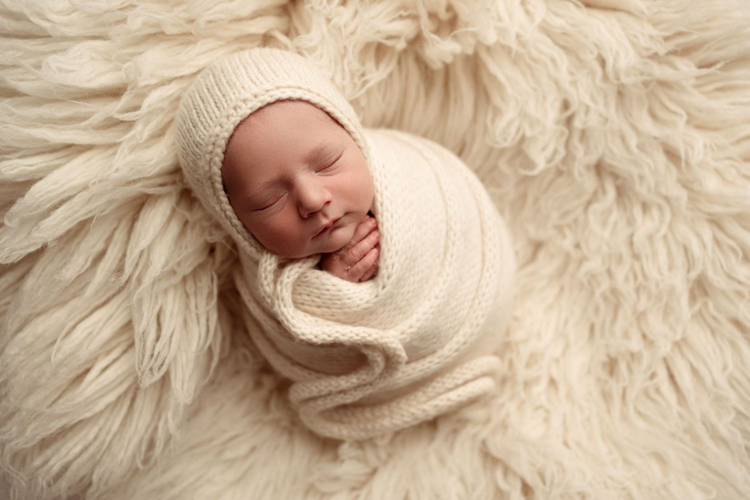 A sleeping newborn wrapped in cozy beige knit blanket rests peacefully in a wooden bowl against rustic background.