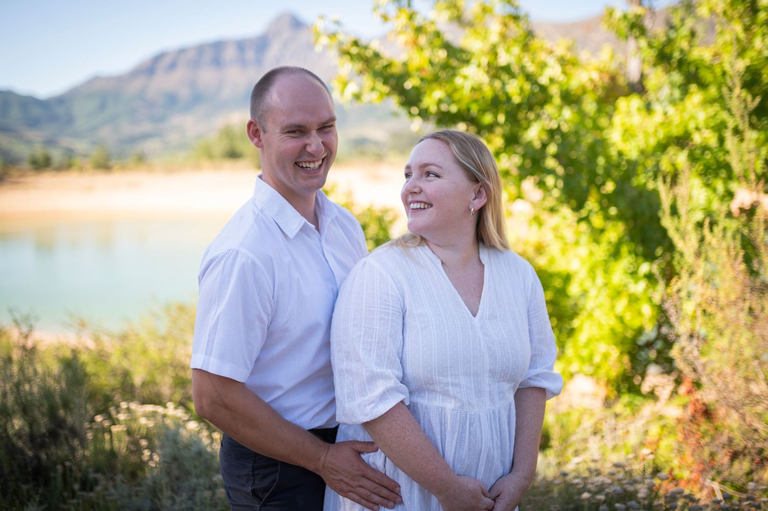 A couple in white shirts smiles at each other by a scenic mountain lake on a sunny summer day.