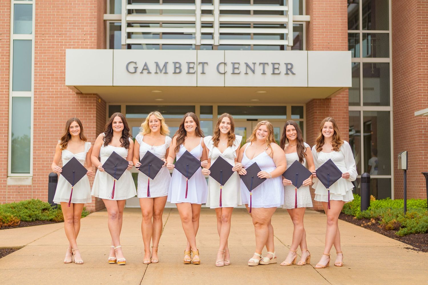 Row of graduates in white dresses outside Gambet Center building.