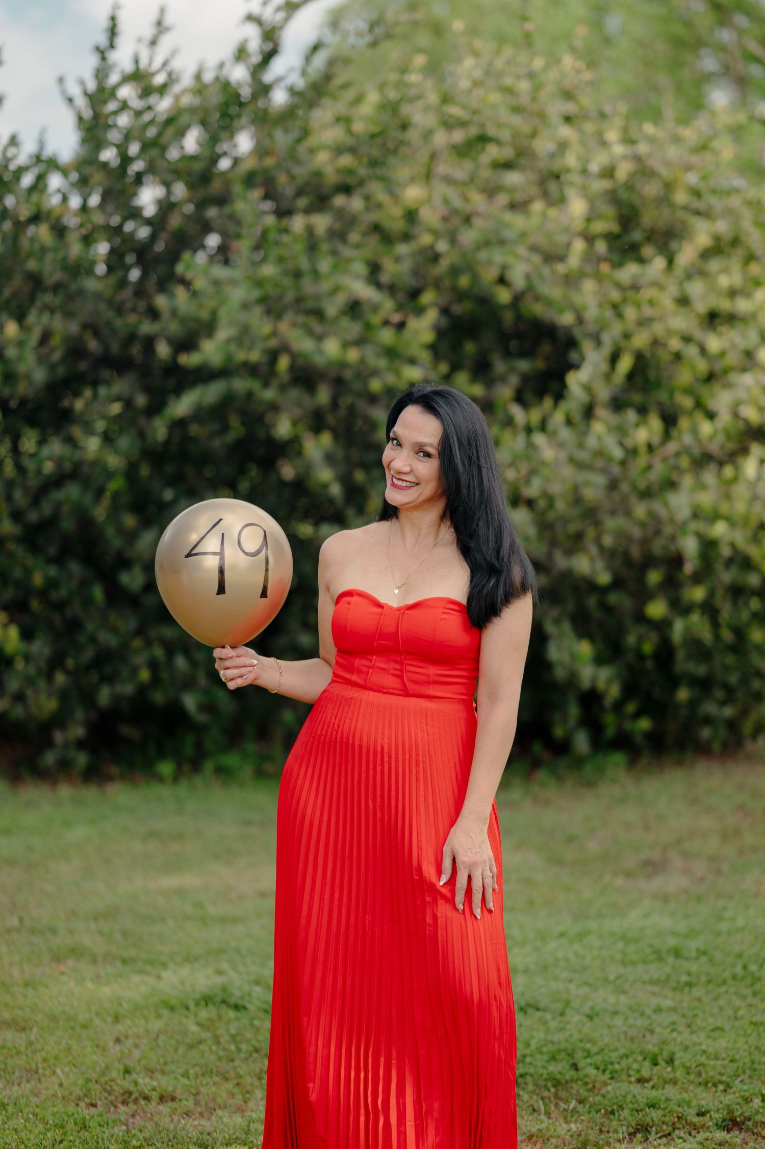 A person in a red pleated gown holds a gold balloon with the number 49 while standing in a garden setting.