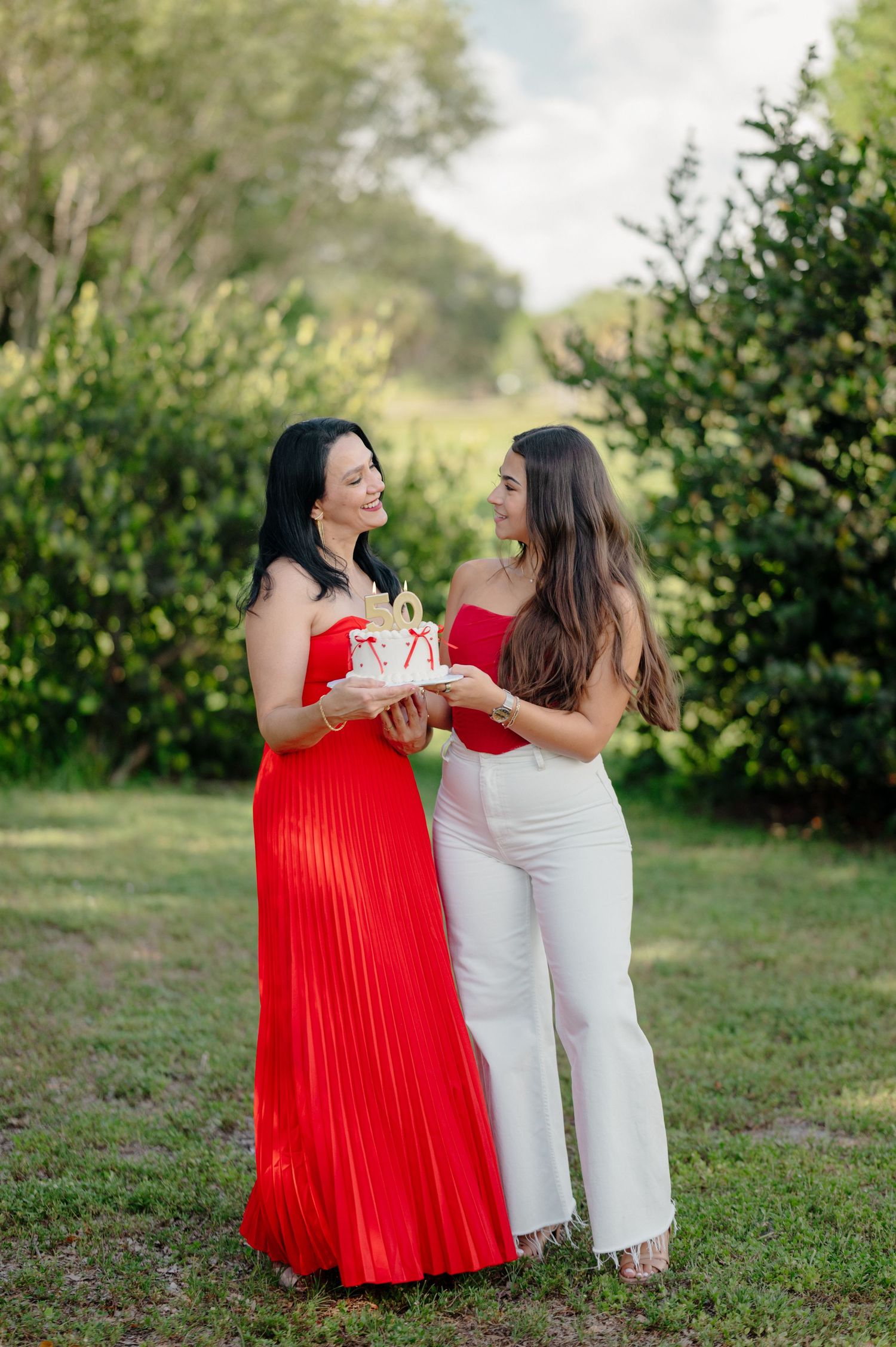 Two women share a joyful moment outdoors, one wearing a flowing red pleated dress and another in white pants.