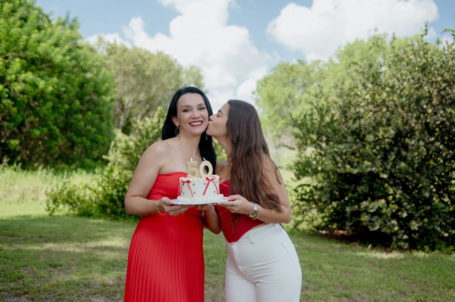 Two women in contrasting red and white dresses share a celebratory moment together in a lush garden setting.
