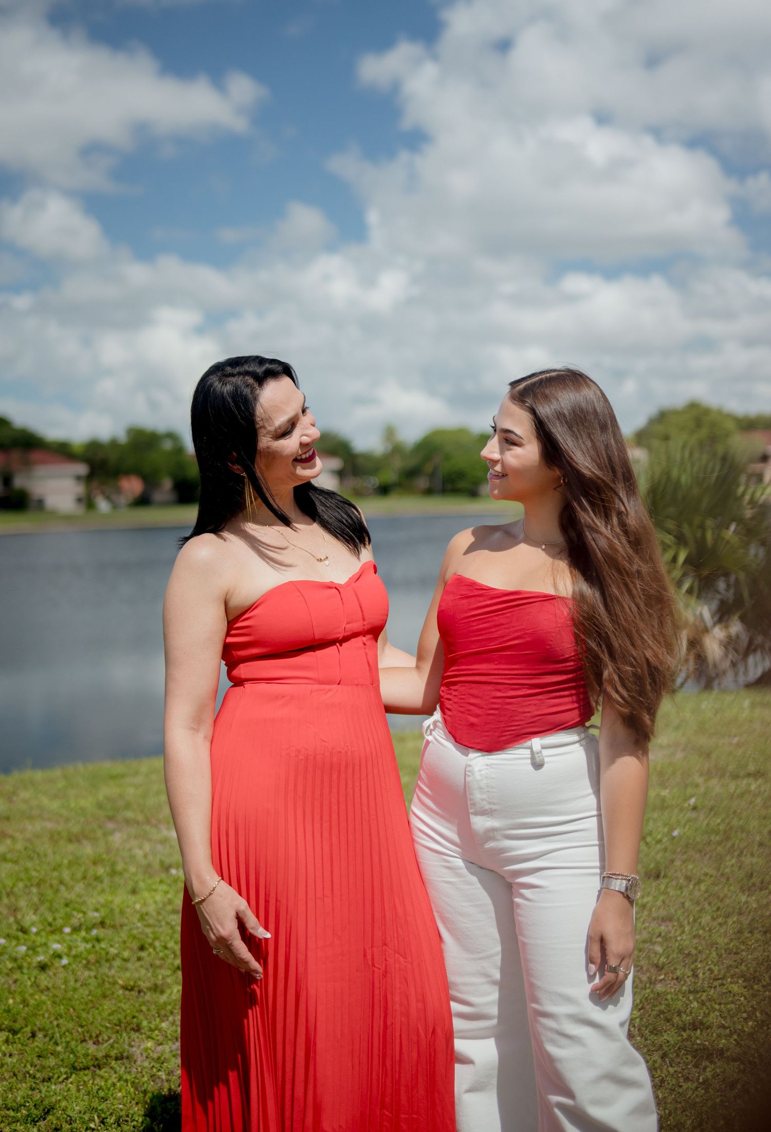 Two people in complementary red and white outfits stand together by a lake under cloudy skies while laughing together.