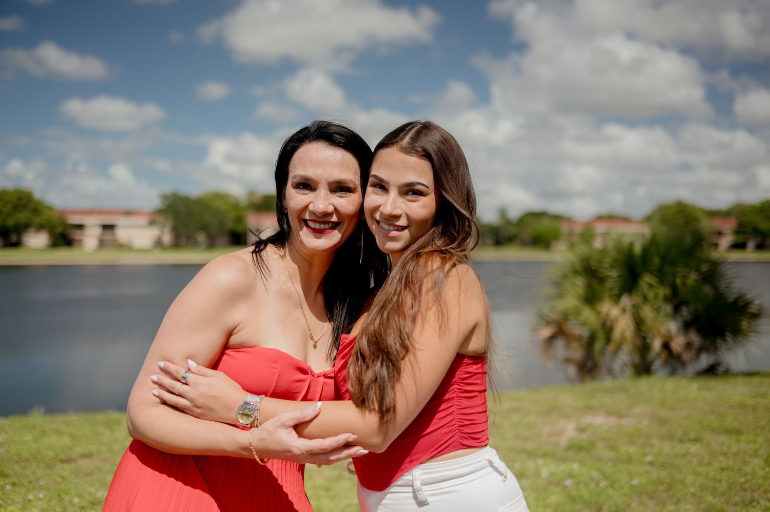 People wearing coral-colored dresses pose together outdoors next to a lake on a sunny day.