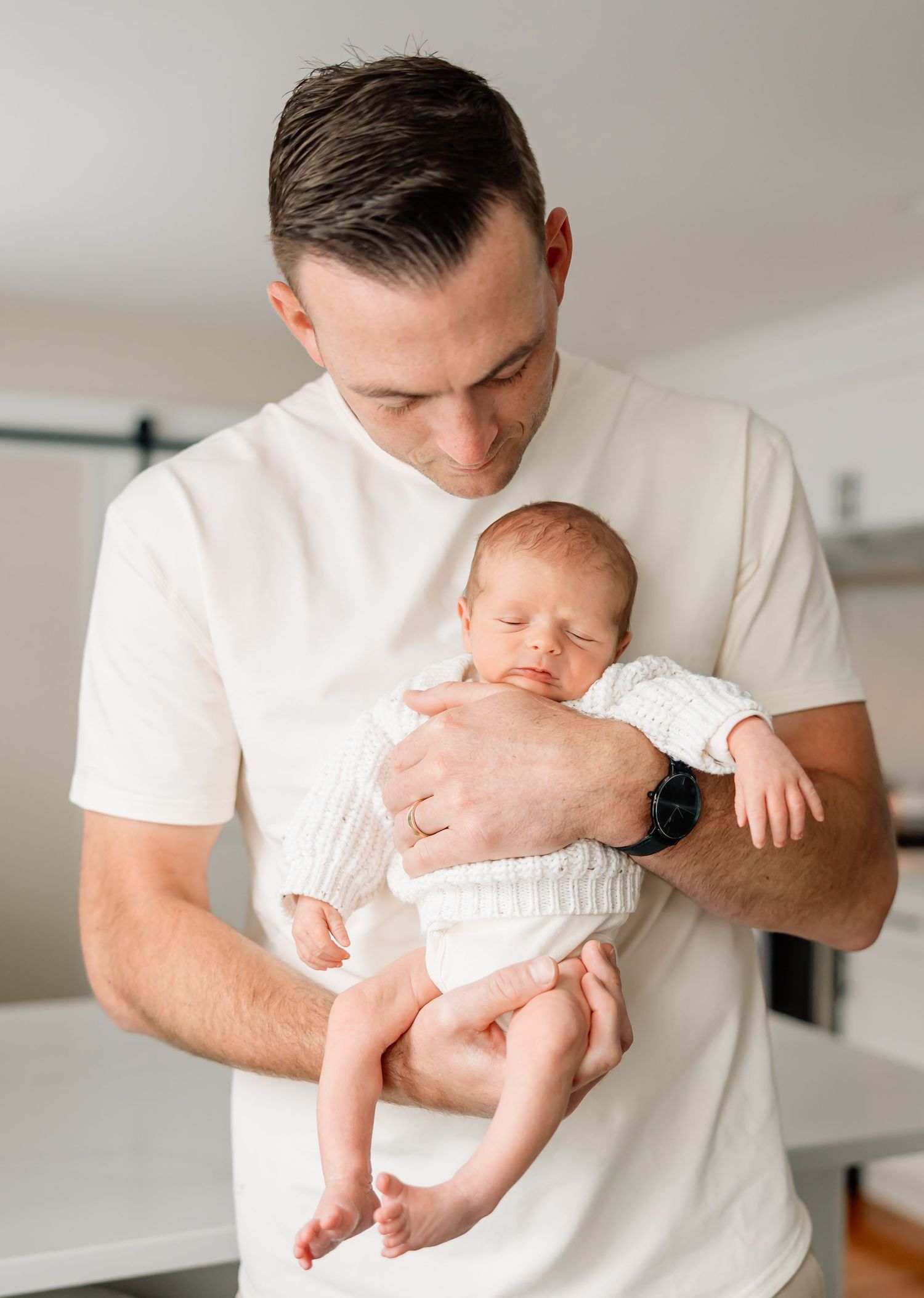 A parent in white clothing holds and kisses a sleeping baby dressed in white against a bright interior backdrop.