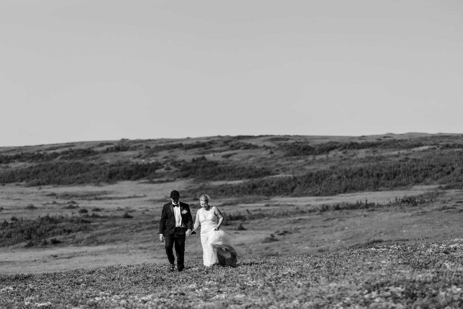 A couple in wedding attire walk hand in hand across a rolling hillside landscape in black and white.