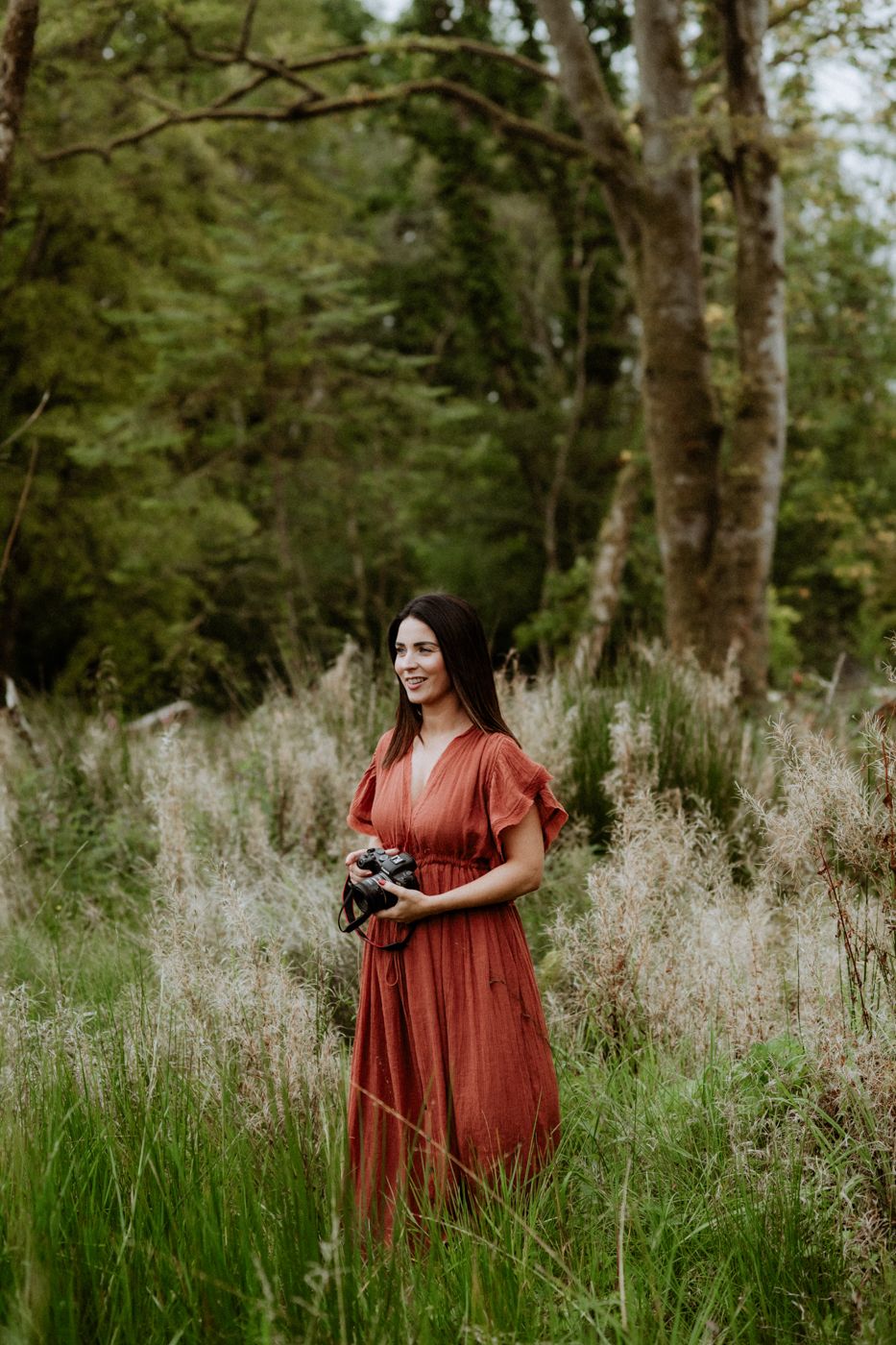 A person in a flowing rust-colored dress stands amidst tall grass and wildflowers in a tranquil, green forest setting, holding a camera.