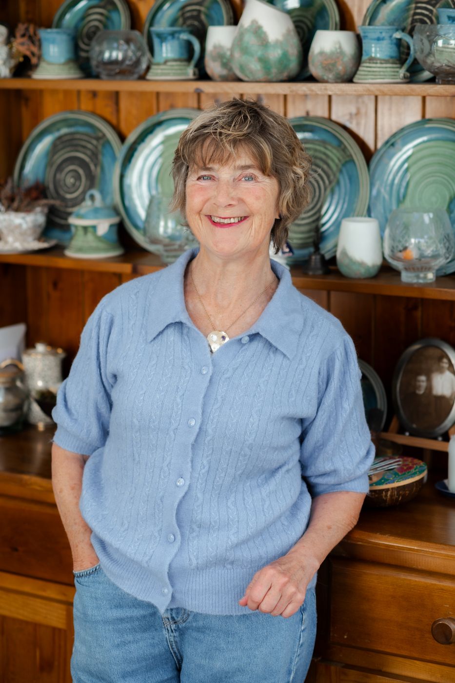 A woman in a light blue cardigan stands confidently in front of a wooden shelf filled with decorative pottery and dishware.