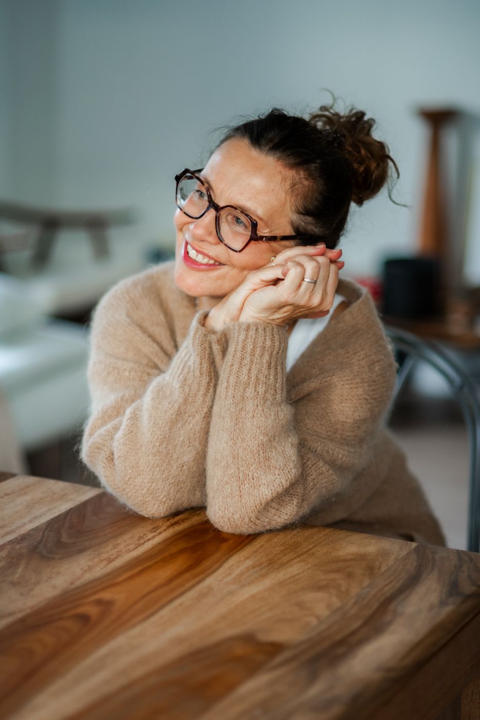 A person rests their chin on crossed arms while sitting at a wooden table, dressed in a cozy beige sweater. Soft, blurred background.