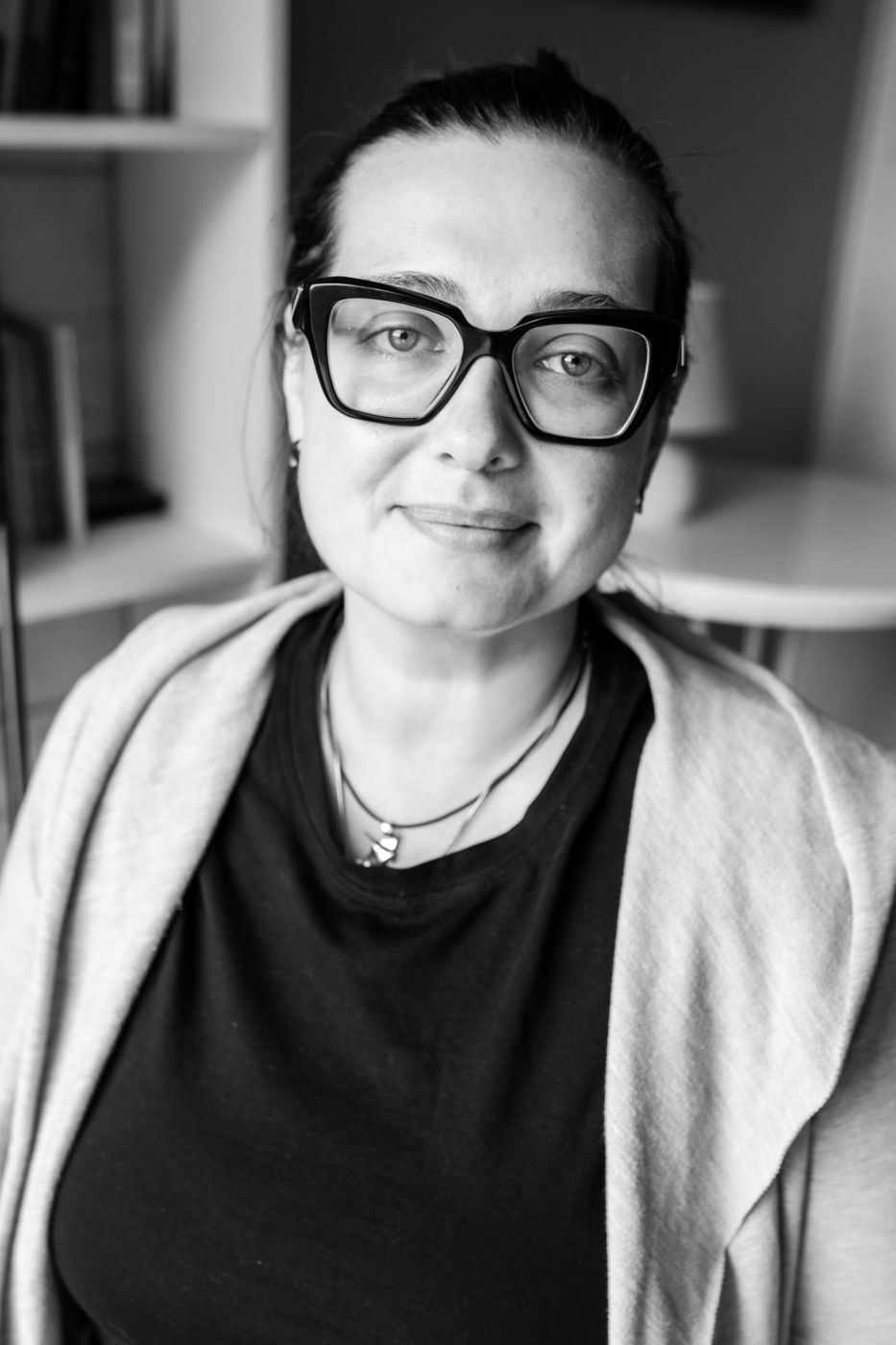 A person wearing a black shirt with layered necklaces sits against a softly blurred background in a monochrome image.