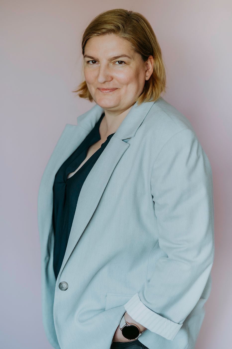 A person stands confidently wearing a light grey blazer over a black shirt, with hands in pockets, set against a soft pink background.
