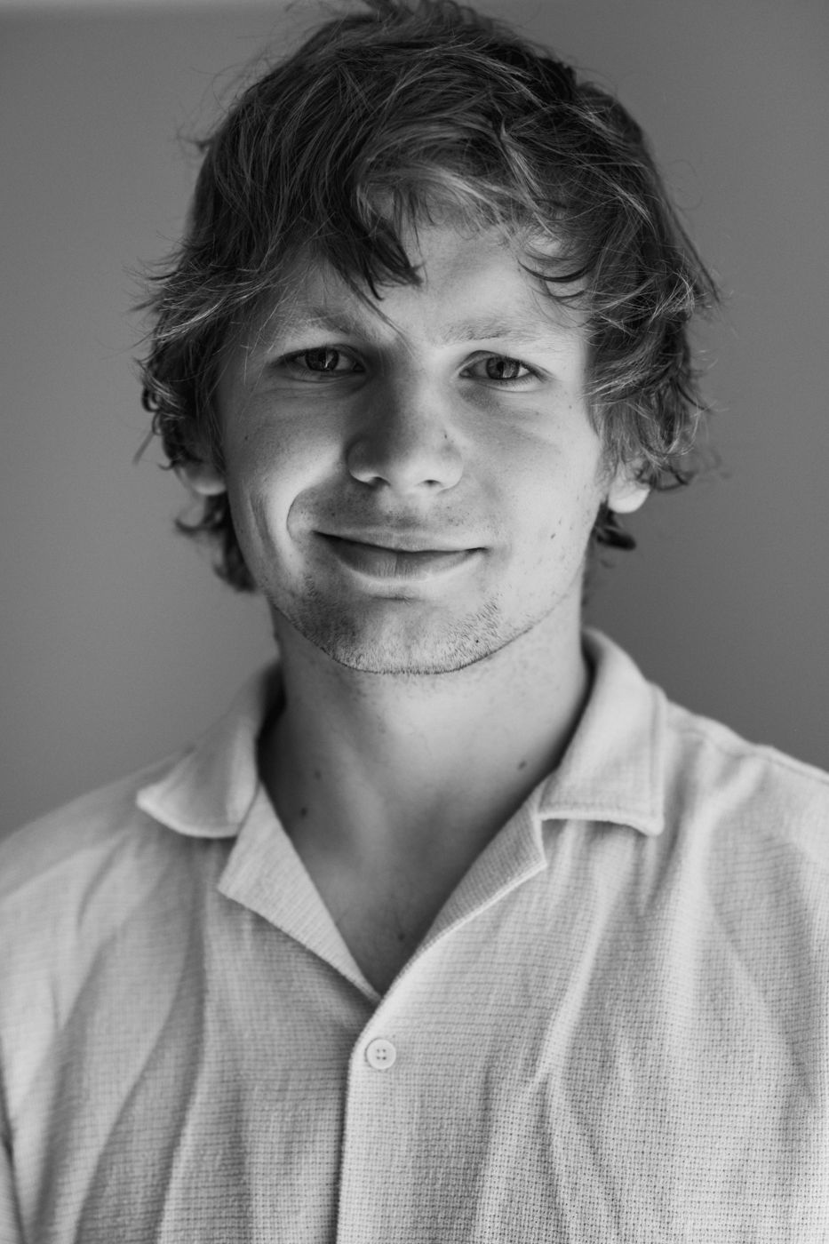 A person with curly hair wearing a light-colored button-up shirt, standing against a soft, neutral background in black and white.