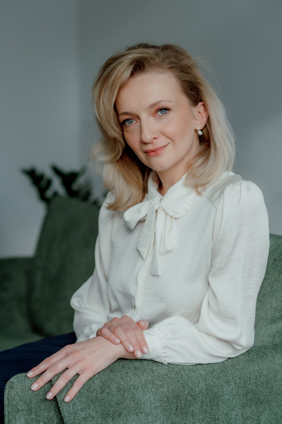 A woman with shoulder-length blonde hair sits on a green couch, wearing a white blouse with a bow tie, hands folded elegantly.