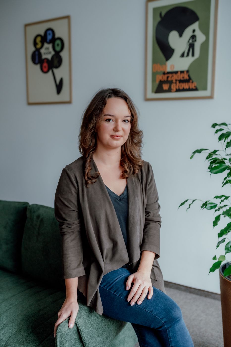 A woman with wavy brown hair sits casually on a green couch, wearing a dark top and a brown jacket, surrounded by indoor plants and artwork.