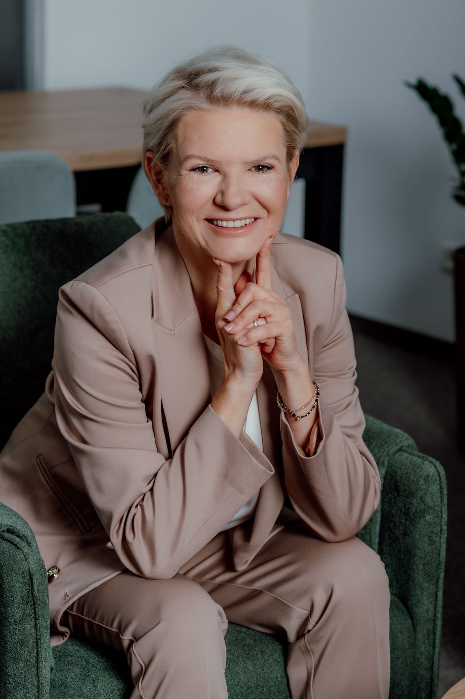 A professionally dressed woman sits on a green armchair, fingers pressed together thoughtfully in a modern office setting.