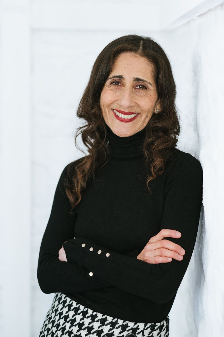 A woman with long curly hair leans against a white wall, wearing a black turtleneck and a patterned skirt, arms crossed confidently.