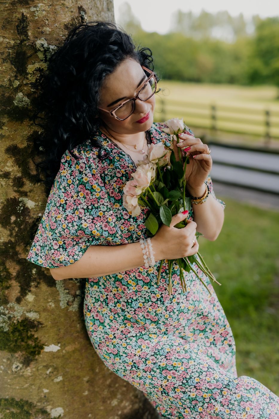 A woman with dark, curly hair and glasses leans gently against a tree, holding a bouquet of soft pink roses close to her chest. Portrait.