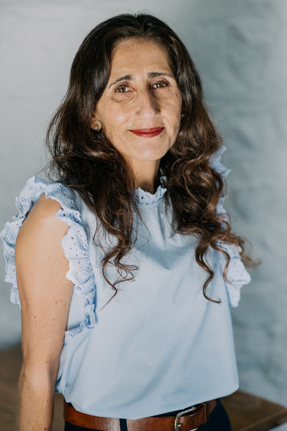 A woman with long, wavy hair wears a light blue blouse with lace ruffles, standing against a softly lit, neutral background.