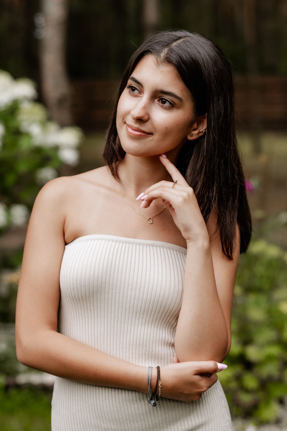 A woman in a fitted beige strapless dress stands outdoors, gently touching her chin, surrounded by lush green foliage and flowers.