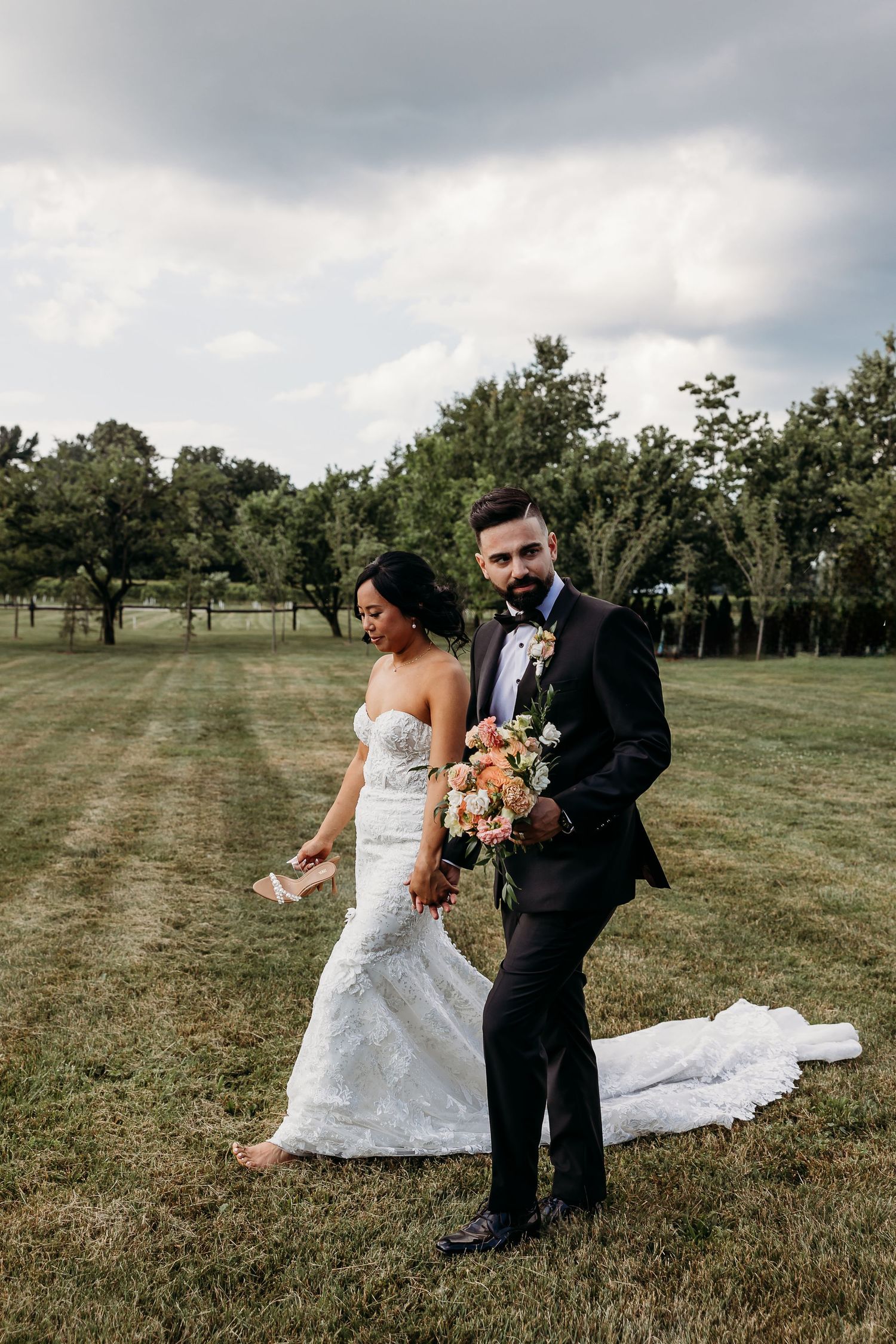 Outdoor wedding photo on grassy field with overcast sky and floral bouquet.