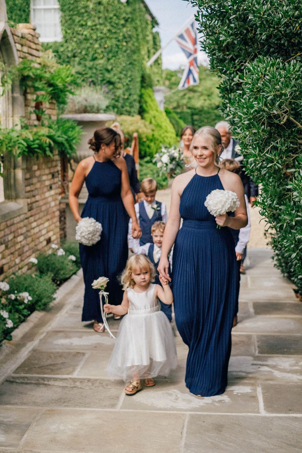 Wedding party in navy dresses walking with flower girl in white along stone path by ivy-covered walls.