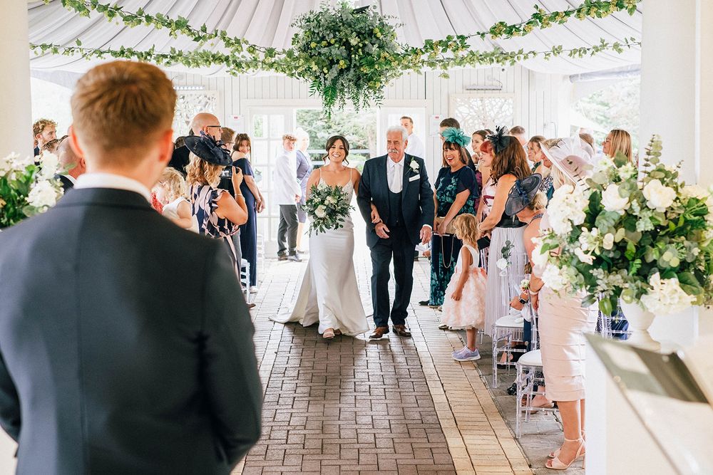 Wedding ceremony in white decorated marquee with guests seated along aisle.