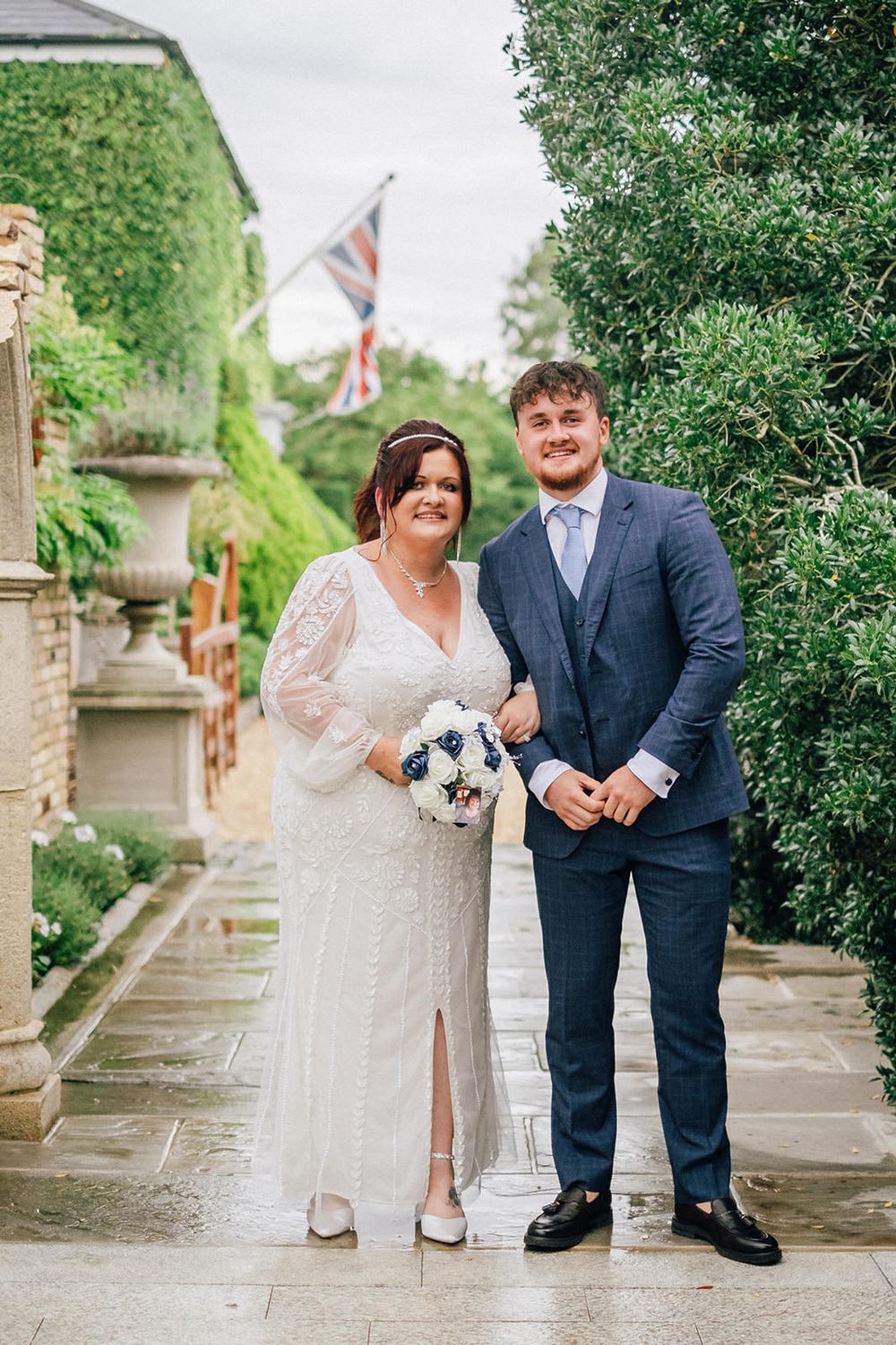 A wedding couple poses together on stone path with Union Jack flag and ivy-covered walls in background.