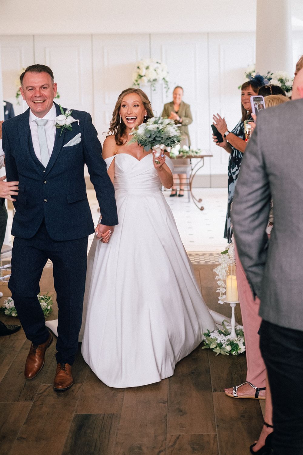 Wedding couple exits ceremony room with joyful expressions as guests celebrate.