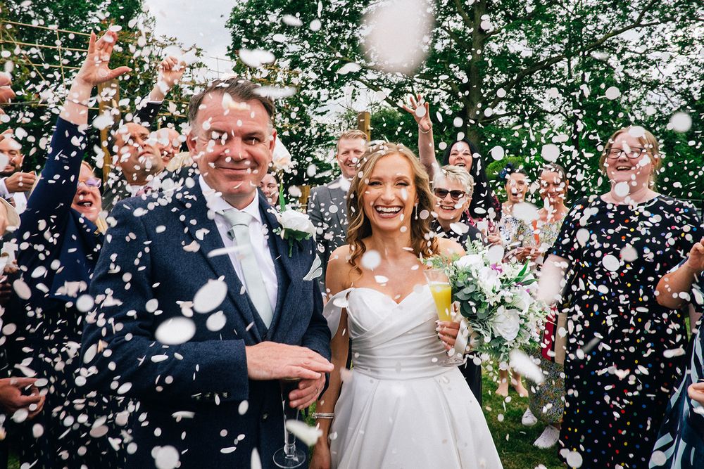 Wedding couple celebrates under shower of white confetti outdoors during ceremony.