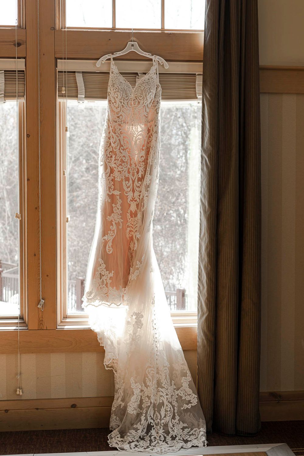 Delicate lace wedding gown hanging in window with natural backlight.