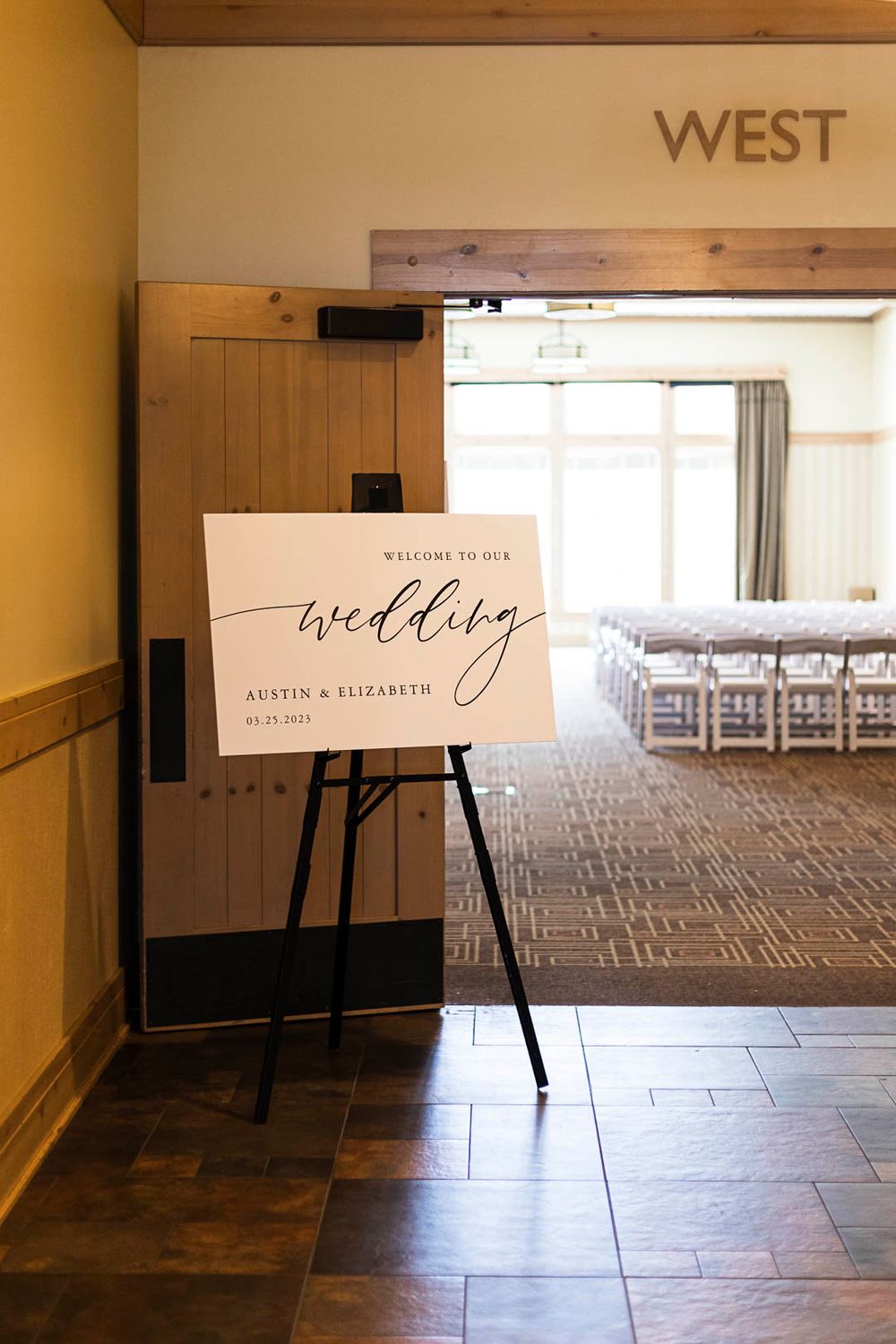 Wedding welcome sign on an easel stands in a hallway entrance to a reception room with white chairs.