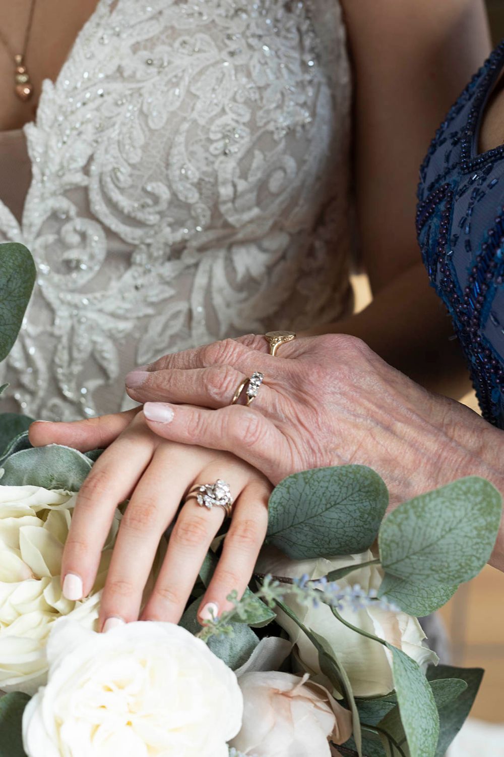 Two hands adorned with elegant rings touch delicately among white roses and eucalyptus leaves.