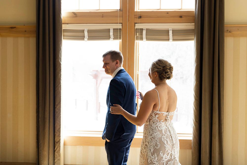 A couple in wedding attire stands by a sunlit window for their first look moment.