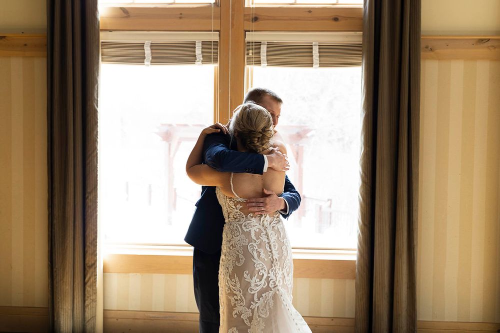 A couple embraces in front of a bright window during an emotional first look.