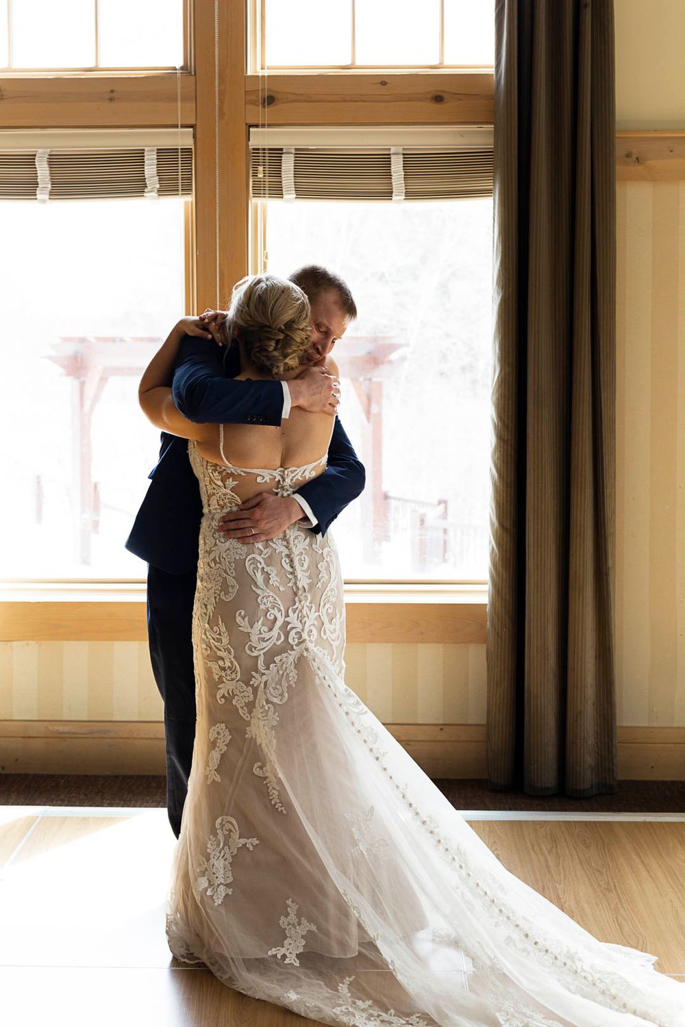 A beautiful wedding embrace captured in front of large windows with flowing lace dress details.