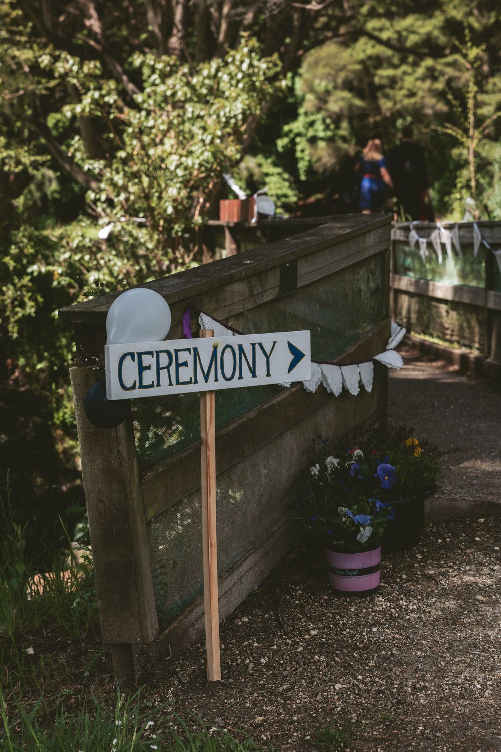 White directional sign reading 'CEREMONY' with arrow mounted on wooden railing in garden setting.