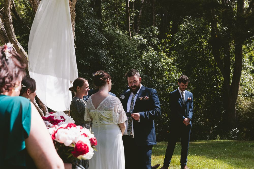 Intimate outdoor wedding ceremony with white draped fabric and red floral arrangements in a wooded setting.