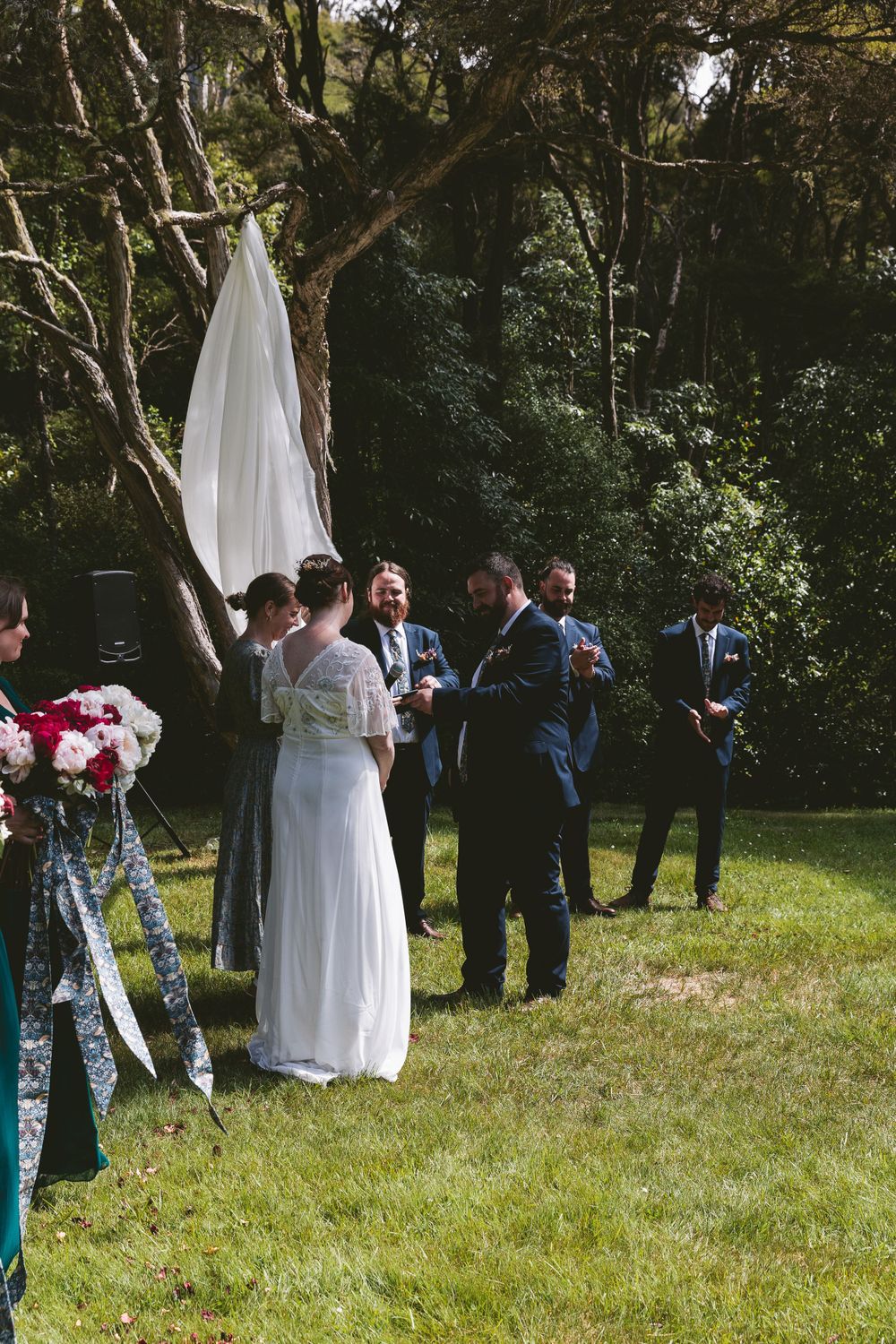 Wedding ceremony in progress under trees with white fabric draping and floral decorations.
