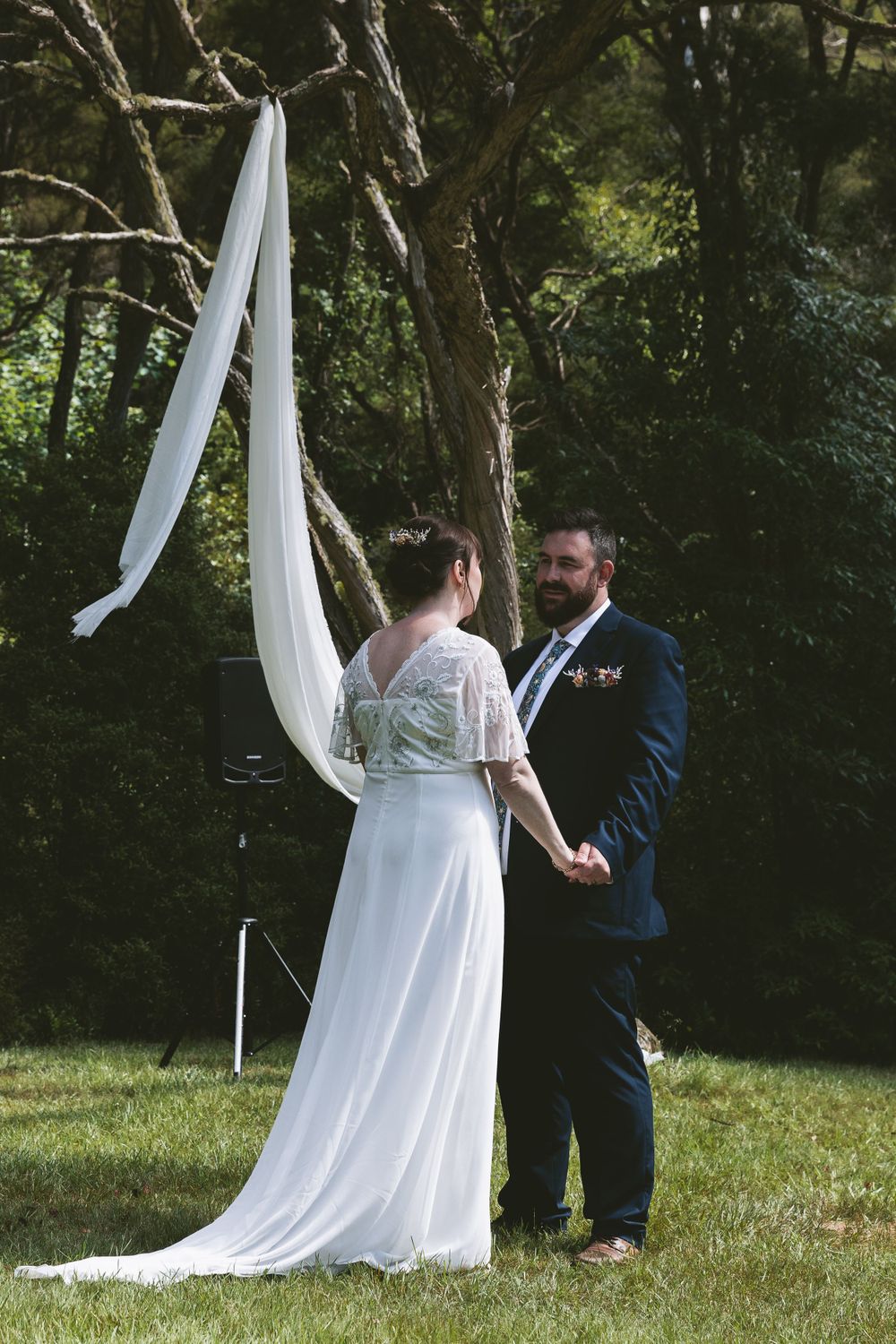 Couple holds hands during wedding ceremony beneath white fabric draped from forest trees.