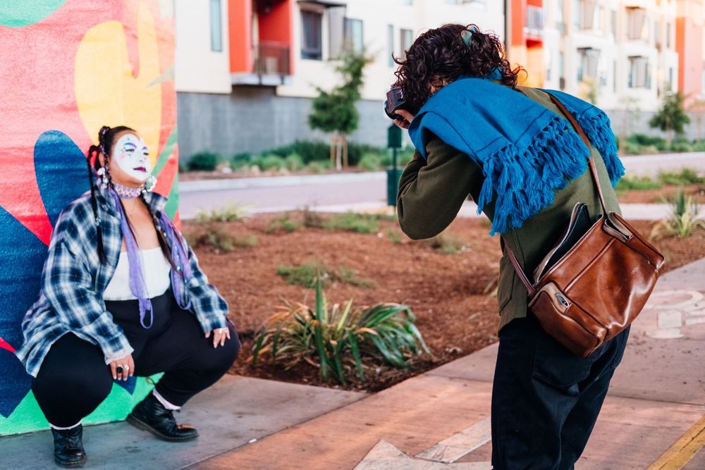 Two people outdoors take photos of each other near colorful buildings, with one person squatting and one bending over.