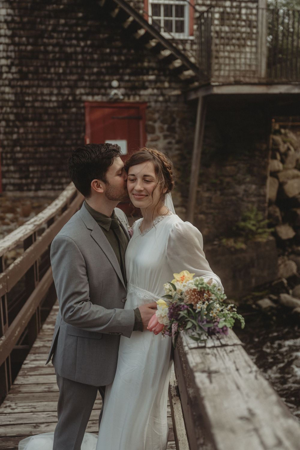 A tender moment shared on a rustic wooden boardwalk near an old brick mill building.