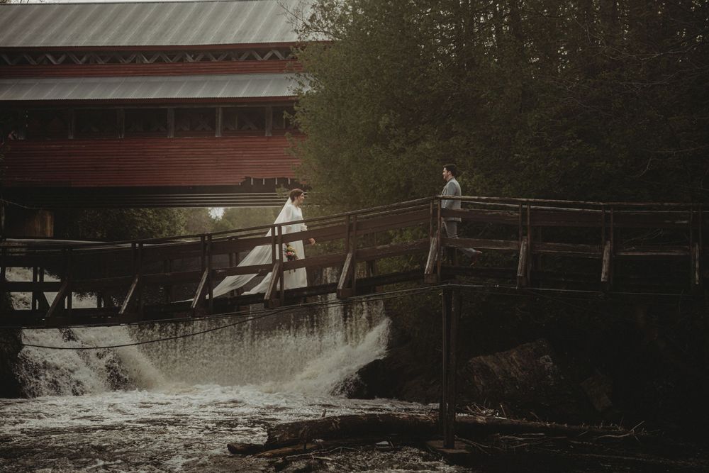 A scenic wedding photo on a wooden bridge over rushing water near historic mill buildings.