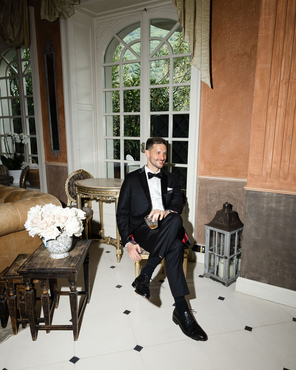 Elegant formal setting with a groom in tuxedo seated in a classic interior with arched windows and decorative lanterns.