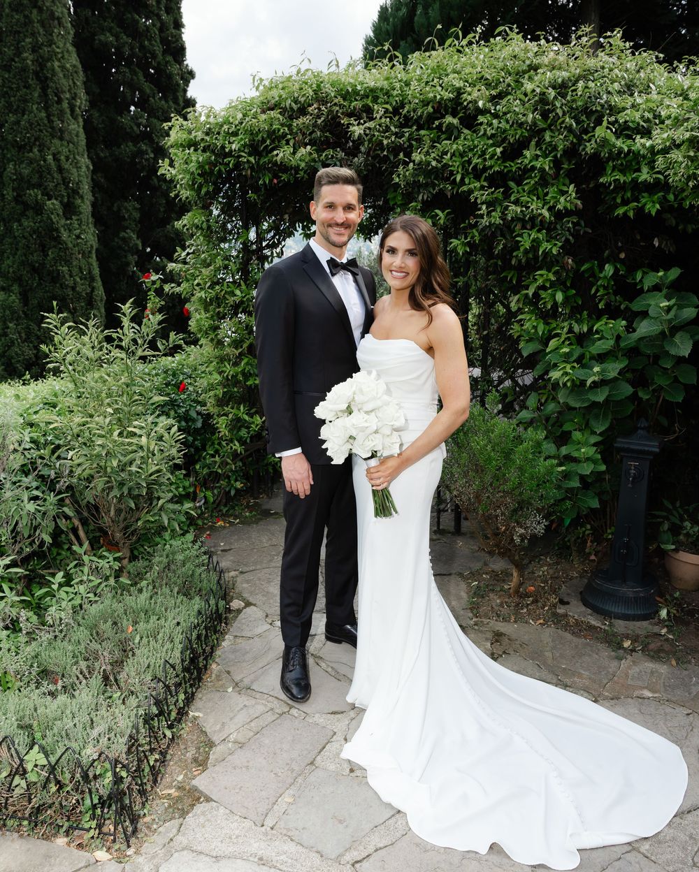 A couple in formal wedding attire poses on a garden path with greenery and cypress trees in the background.